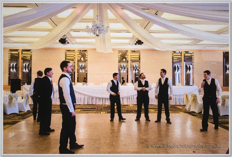 Groomsmen in vests scatter across the draped Grand River Ballroom beneath a crystal chandelier