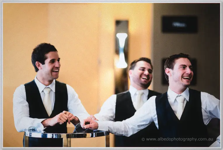 Three groomsmen lean on a foyer table laughing heartily in soft golden light