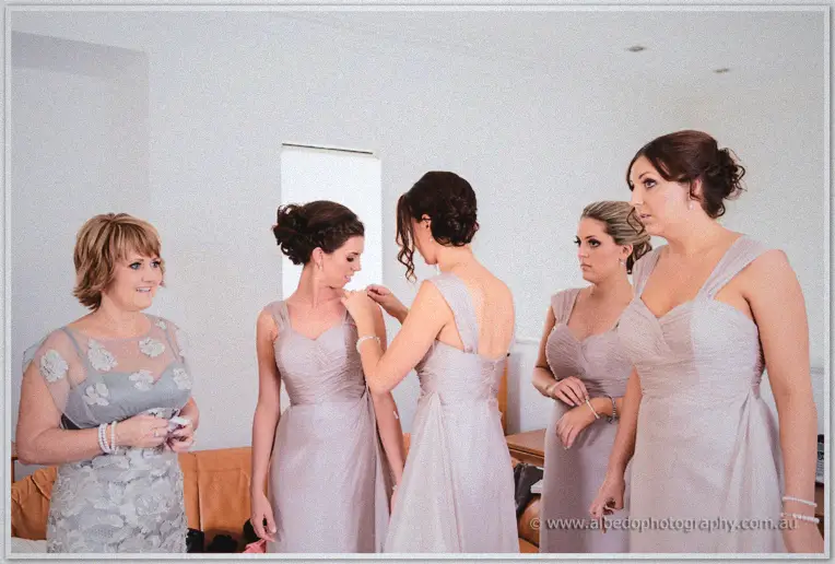 Bridesmaids in dusty lilac gowns help fasten a necklace in a bright morning room