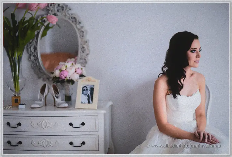 Karis sits beside a mirrored dresser holding her shoes, bouquet and a framed picture