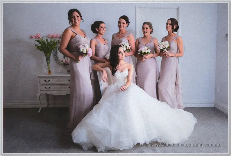Bridesmaids in taupe gowns stand behind Karis as she sits in her full white gown