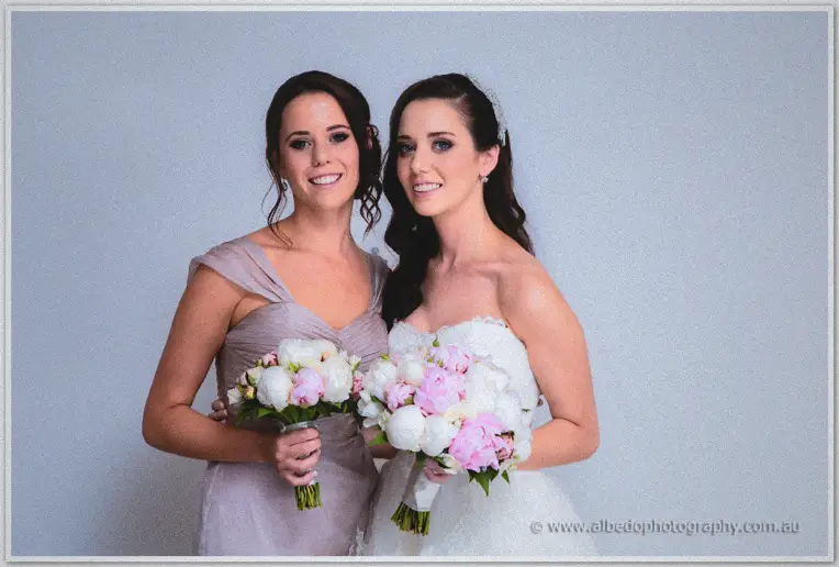 Bride and bridesmaid smile side by side holding pink and white peony bouquets