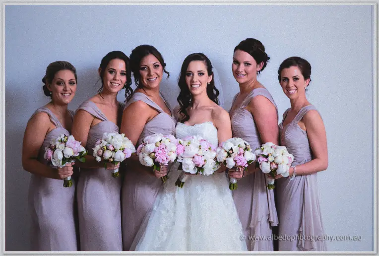 Karis and five bridesmaids line up holding pink and white peony bouquets against a grey wall