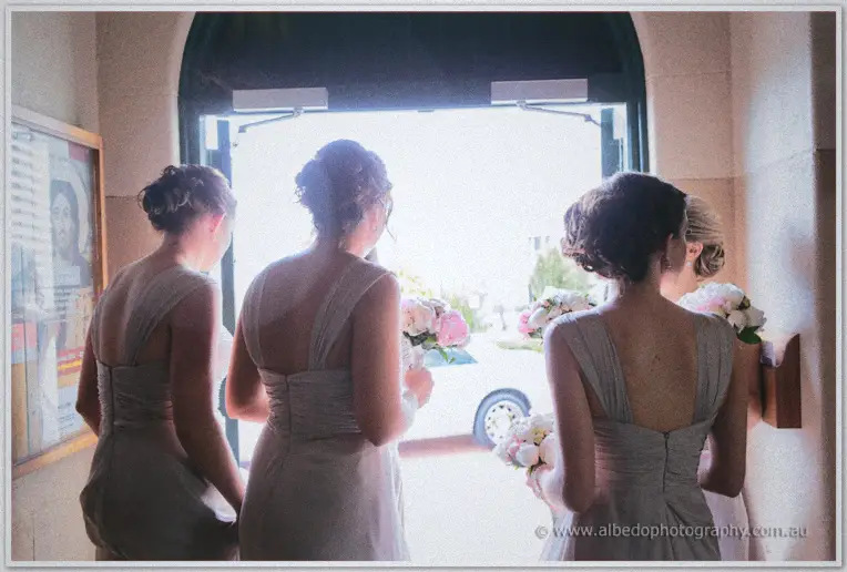 Bridesmaids face the bright church doorway holding bouquets under an arched entry