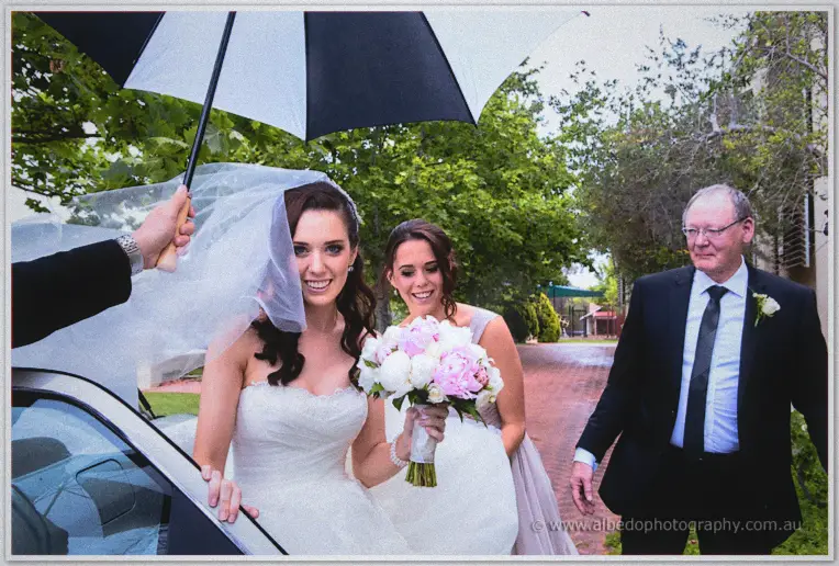 Karis steps out in drizzle as an umbrella shields her veil and her father walks close