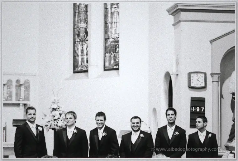 Black and white line of groomsmen in tuxedos waiting near stained glass windows