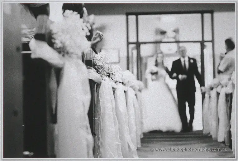 Karis walks the flower-lined aisle on her father's arm in a soft black and white frame