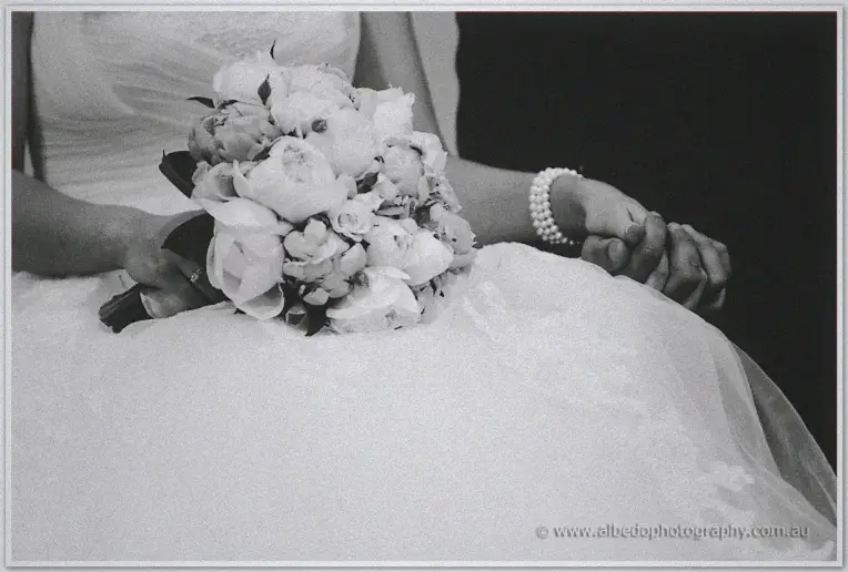 Karis holds her peony bouquet in her lap while hands clasp beside a pearl bracelet