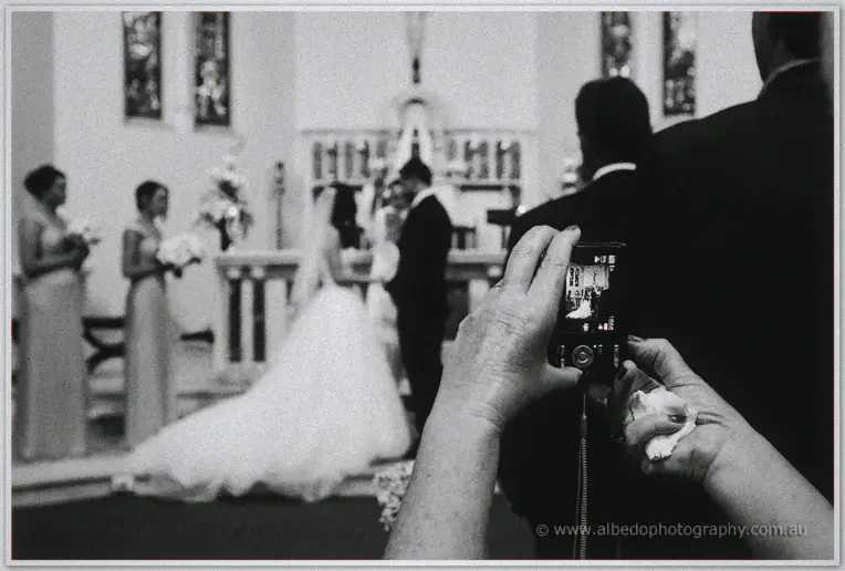 A guest raises a compact camera over the pews as Karis and Nathan marry at the altar