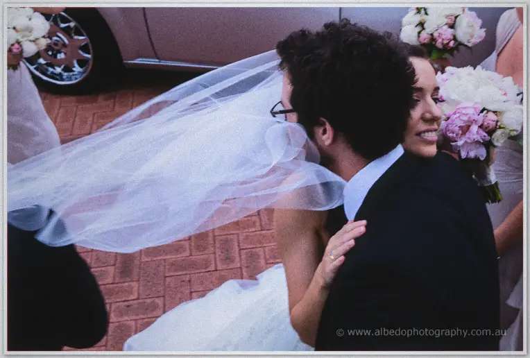 Karis hugs a guest by the car as wind whips her veil past bridesmaids with peonies