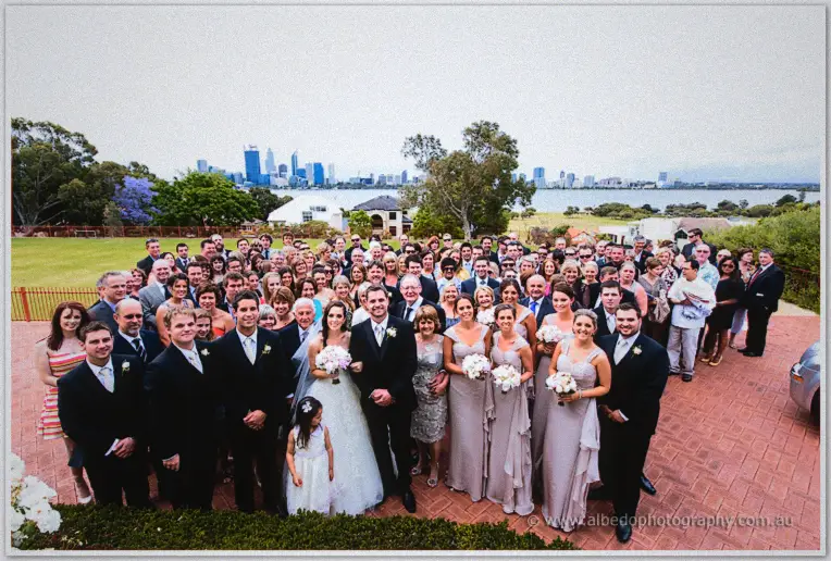 Karis and Nathan pose with their wedding guests on a brick terrace beneath the Perth skyline and river