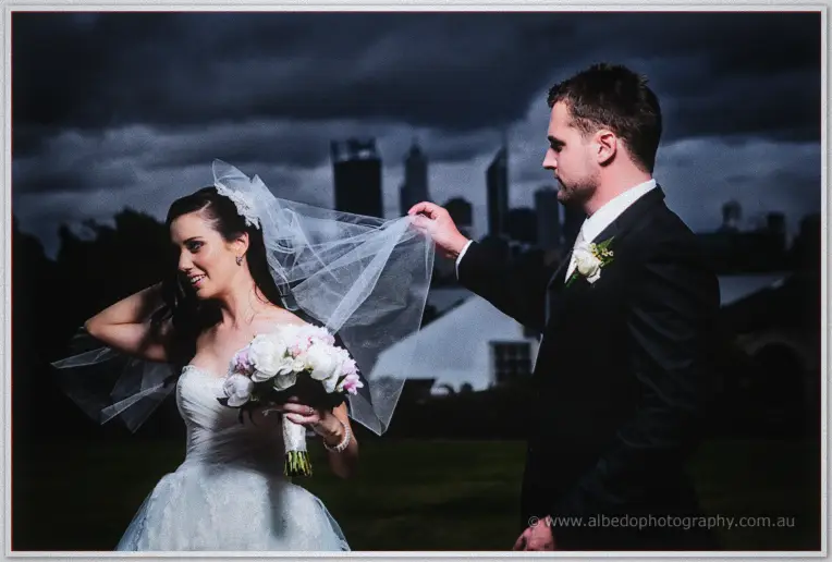Nathan lifts Karis's billowing veil against a moody storm sky with the Perth skyline behind them