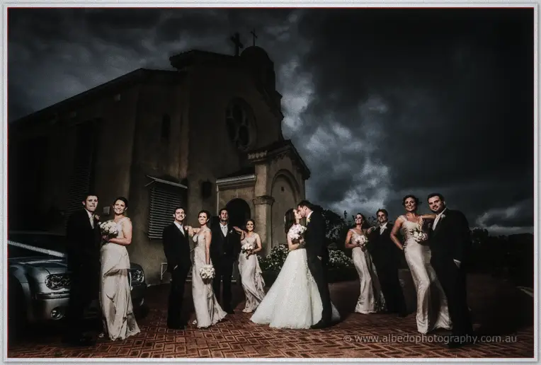 The bridal party flanks Karis and Nathan kissing before a stone church under darkening storm skies