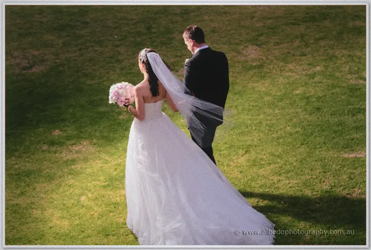 Seen from behind, Karis carries her bouquet beside Nathan as they walk across a sunlit green lawn