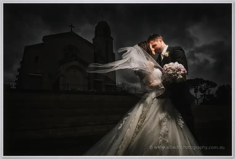 Nathan kisses Karis beneath her windswept veil outside a stone church under stormy skies