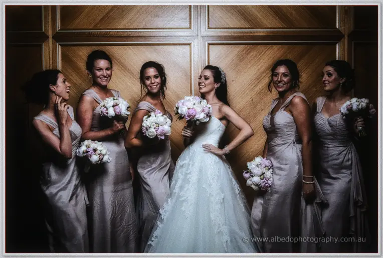 Karis laughs with her six bridesmaids in silver gowns holding peony bouquets against timber panelled walls