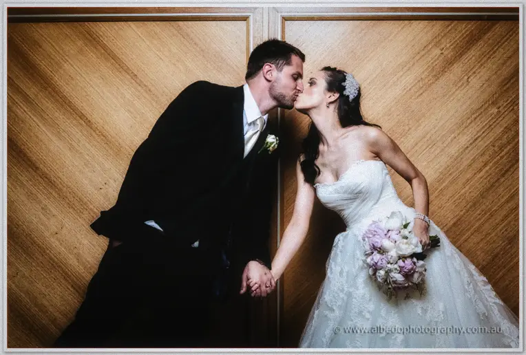 Hand in hand, Karis leans up to kiss Nathan against the ballroom's chevron timber wall