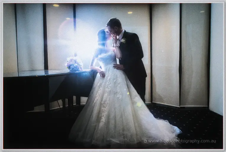 Karis and Nathan kiss beside a grand piano as backlight streams through the curtained windows