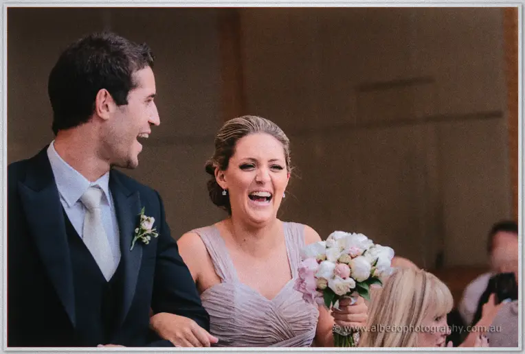 A groomsman and a bridesmaid in taupe double over laughing during the reception speeches