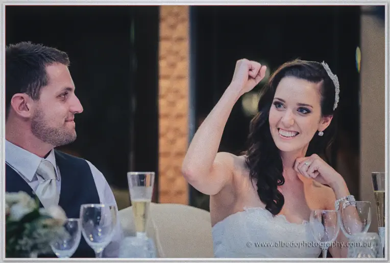 Karis pumps her fist and laughs at the bridal table while Nathan watches over champagne glasses