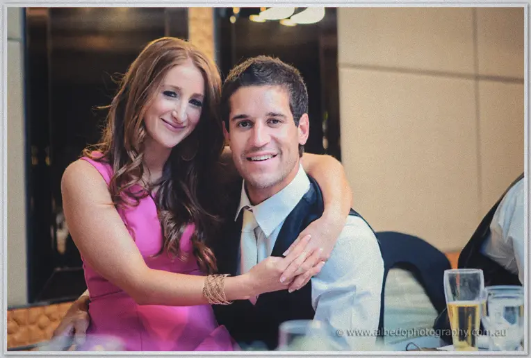 A guest in pink hugs a smiling friend in vest and tie at the reception table