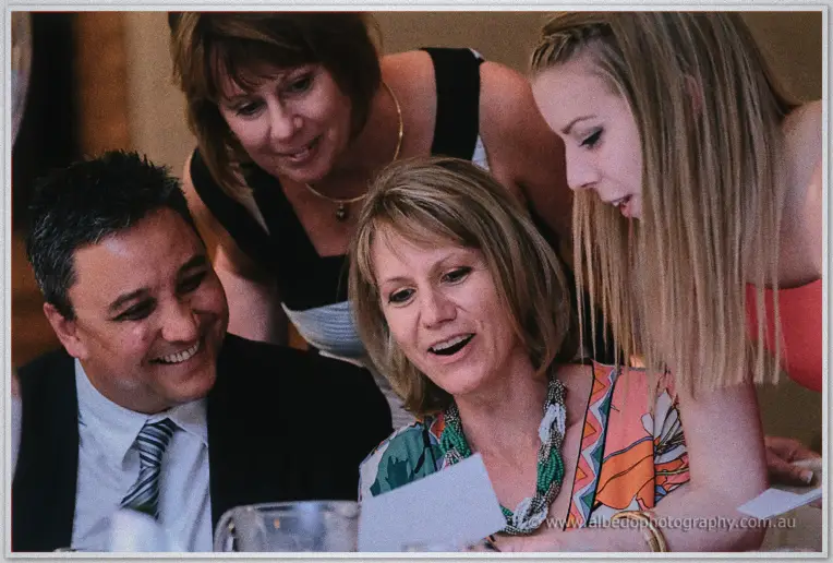 Four guests huddle laughing over a card at their table in the lamplit ballroom