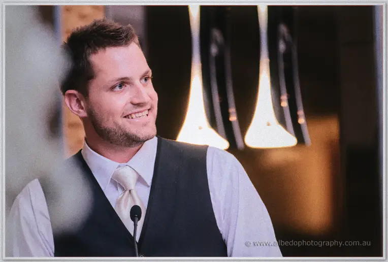 A groomsman grins at the bridal table, pendant lights glowing gold behind him