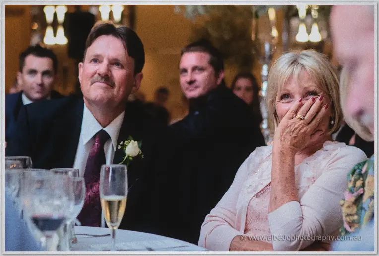 A teary guest covers her smile beside the groom's father in a burgundy tie