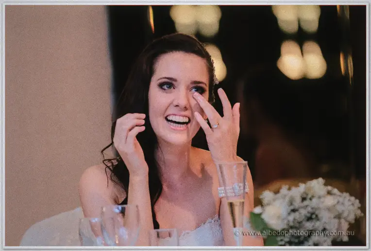 Karis wipes happy tears at the bridal table, chandelier glow and white blooms behind her