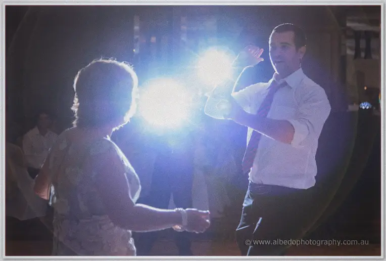 A groomsman in a loosened tie dances theatrically with a wedding guest amid lens flare