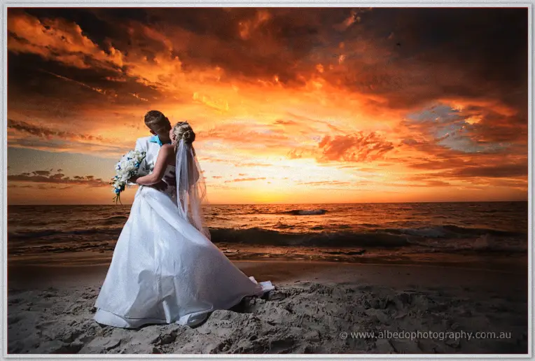Lauren and Guy embrace on the shoreline beneath a fiery orange sunset sky