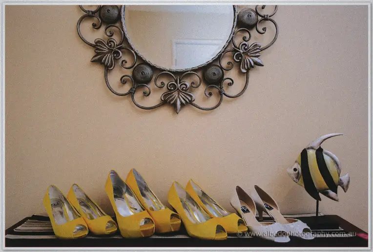 Bright yellow bridesmaid heels and white bridal shoes lined up beneath an ornate wrought iron wall mirror