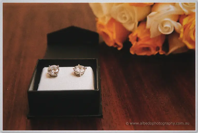 Diamond stud earrings in an open black box beside a bouquet of orange and cream roses on dark timber