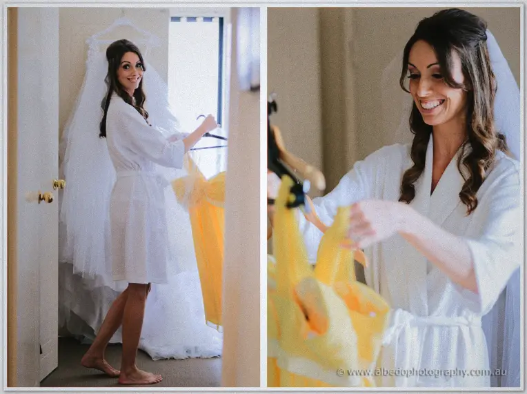 Bride in a white robe smiling beside her hanging veil while holding a yellow bridesmaid dress