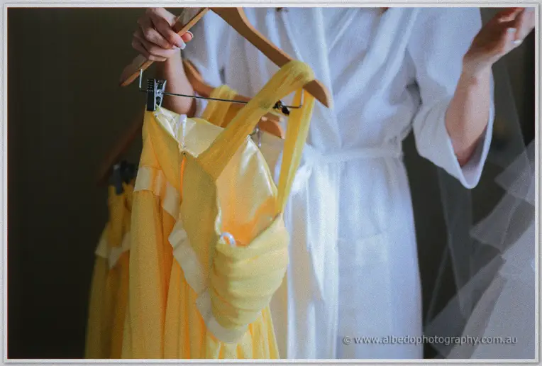 Close-up of sunny yellow bridesmaid dresses on wooden hangers beside a bride in a white robe