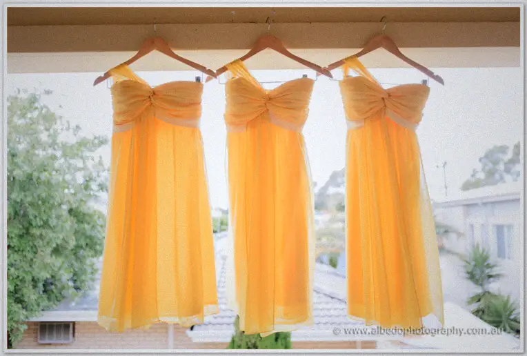 Three yellow one-shoulder bridesmaid dresses hanging on wooden hangers against soft outdoor light