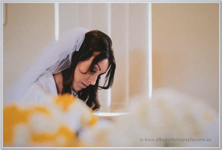 Veiled bride with dark curls gazing down over orange and cream bouquets in warm window light