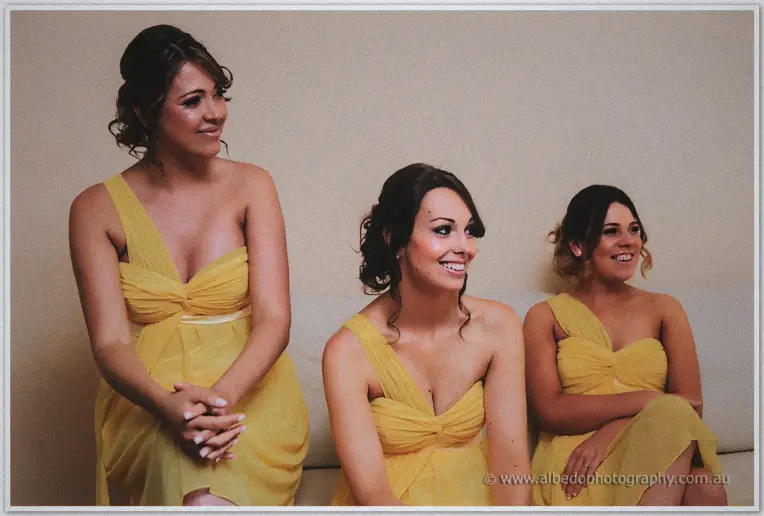 Three bridesmaids in yellow one-shoulder dresses sitting side by side and laughing on a couch