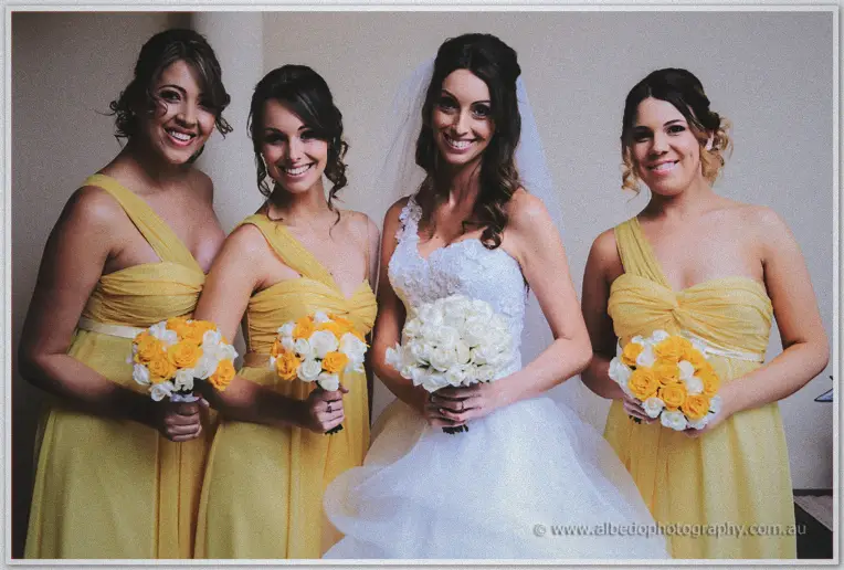 Bride holding white roses beside three bridesmaids in yellow clutching orange posies indoors
