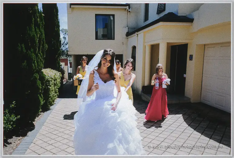 Veiled bride leading her laughing bridal party down a brick driveway past cream townhouses in sunshine