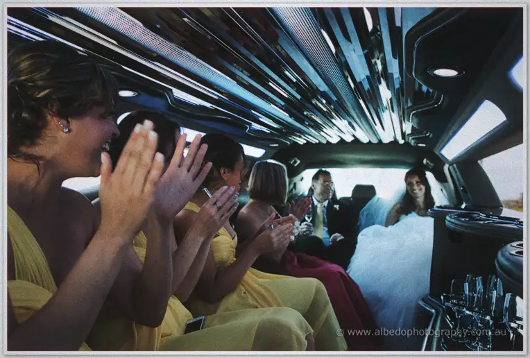Bridesmaids clapping inside a neon-lit limousine while the couple sits at the far end