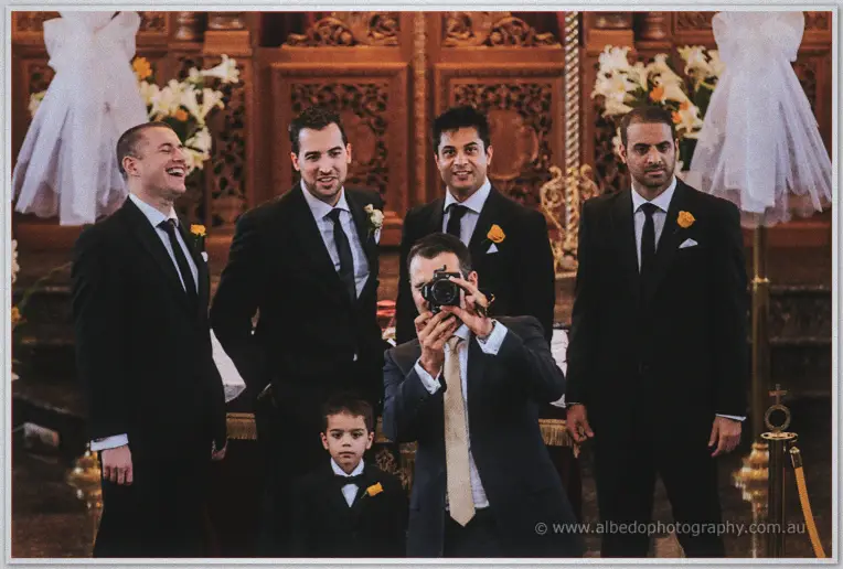 Groomsmen with yellow rose boutonnieres laughing as one aims a vintage camera at the lens