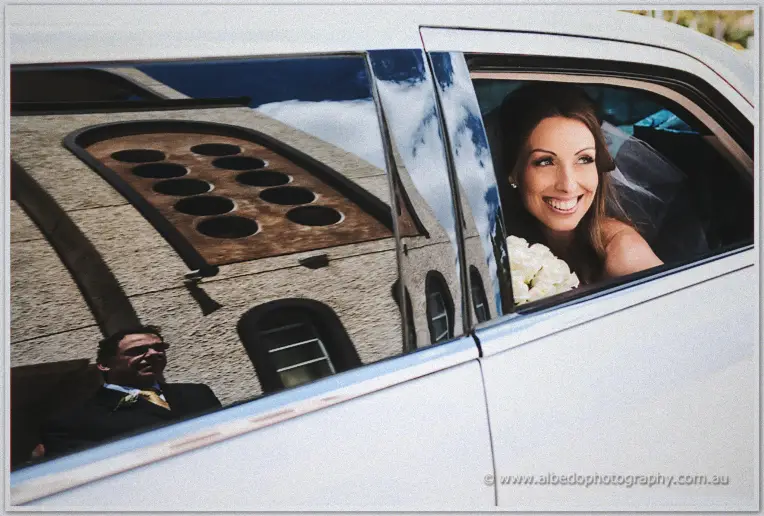 Beaming bride leaning from the limousine window with white roses as the church reflects in the door