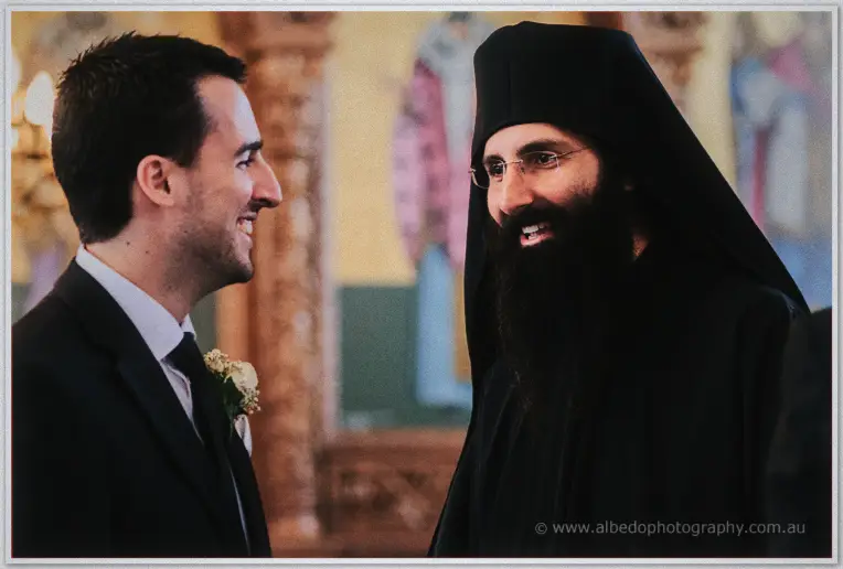 Bearded Greek Orthodox priest sharing a laugh with the groom in his dark suit before the service