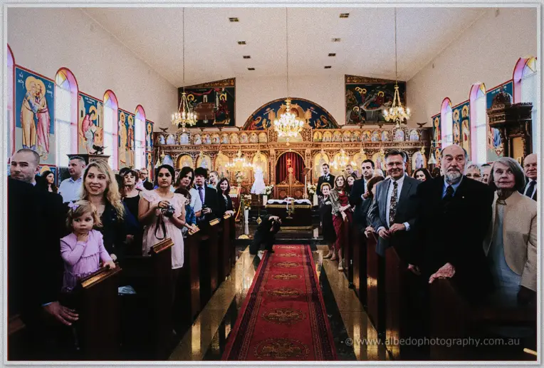 Wedding guests standing among the pews of an icon-filled Orthodox church beneath glowing chandeliers