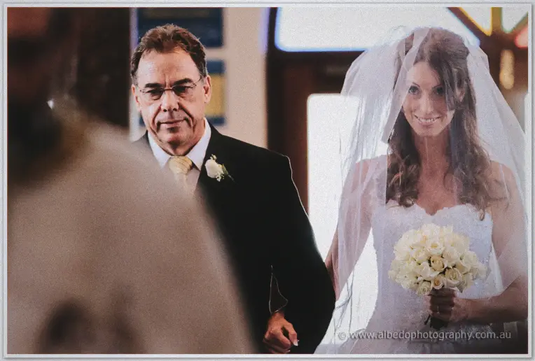 Father walking his veiled bride slowly down the aisle as she smiles holding white roses