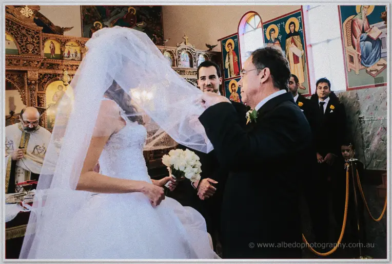 Father lifting the bride's veil beside the groom as the priest looks on at the icon screen