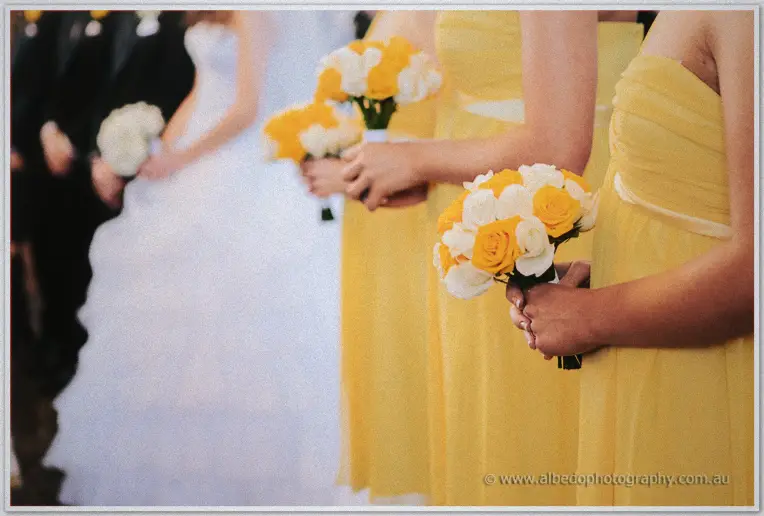 Yellow and white rose bouquets held by bridesmaids as the veiled bride waits behind