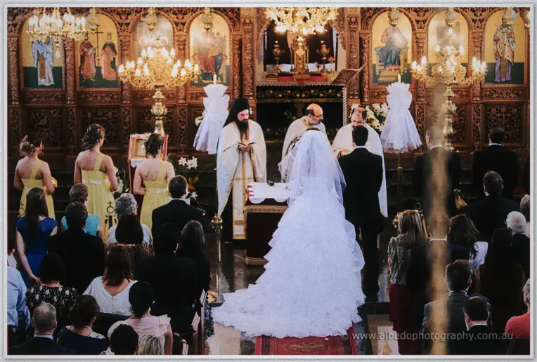 Couple standing before two priests at the candlelit iconostasis while guests watch from the pews
