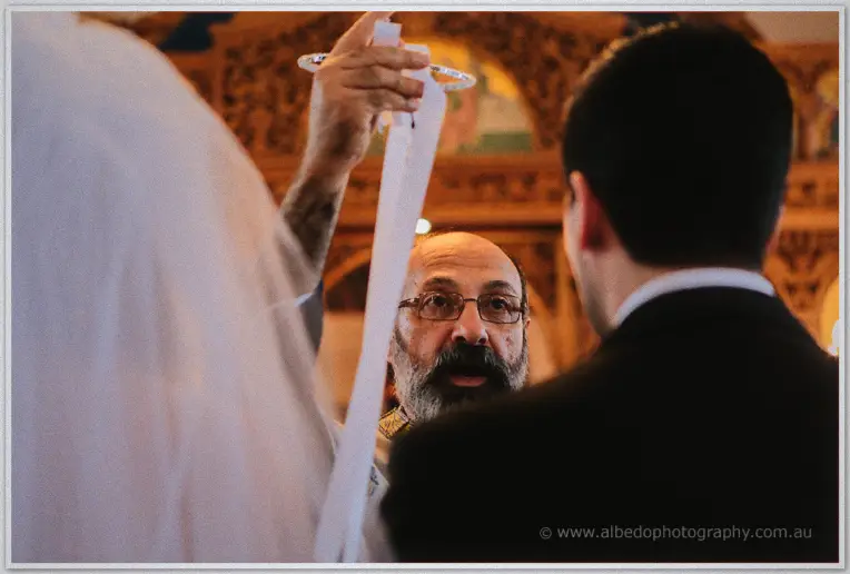 Priest placing ribboned crowns above the bride and groom during their Orthodox wedding ceremony